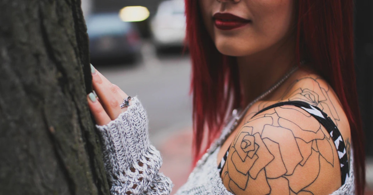 young woman with tattoo on her shoulder, wearing an off-the-shoulder sweater and leaning against a tree