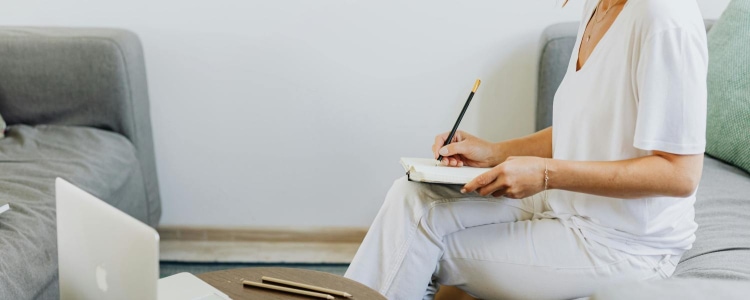 woman wears loose white women's weekend t-shirt with white jeans; she is sitting on a couch and writing in a journal. On a coffee table in front of her there is a laptop, a cup of coffee, and a variety of scissors and writing utensils.