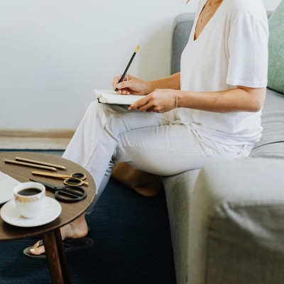 woman wears loose white women's weekend t-shirt with white jeans; she is sitting on a couch and writing in a journal. On a coffee table in front of her there is a laptop, a cup of coffee, and a variety of scissors and writing utensils.