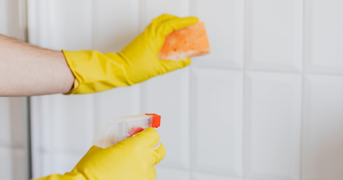 yellow-gloved hands spraying cleaning solution at white tile and scrubbing with an orange sponge