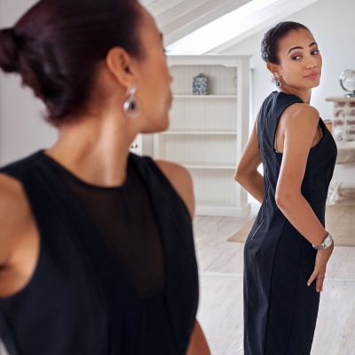 black businesswoman trying on a black sheath dress in a mirror at home