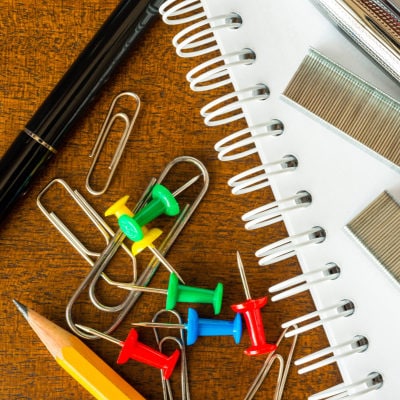 school supplies such as paperclips, pushpins, pencils and pens sit near a white notebook with staples on top