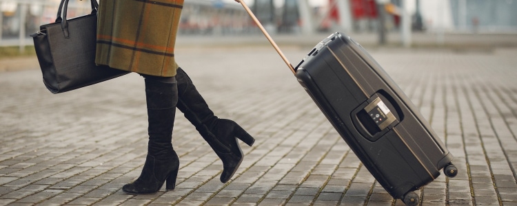 businesswoman wears suede knee-high boots with a work outfit; she is walking outside on cobblestones and pulling a suitcase; she is wearing a brown checkered coat