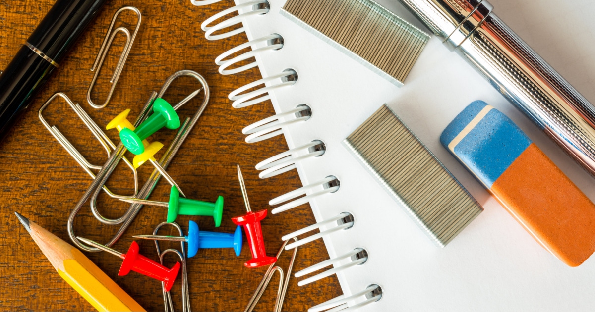school supplies such as paperclips, pushpins, pencils and pens sit near a white notebook with staples on top