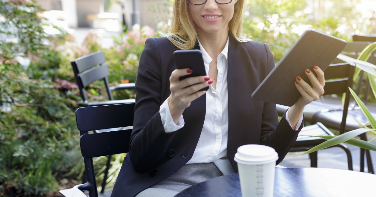 professional young woman looks at her phone while sitting outside; she wears a suit and has a paper coffee cup in front of her