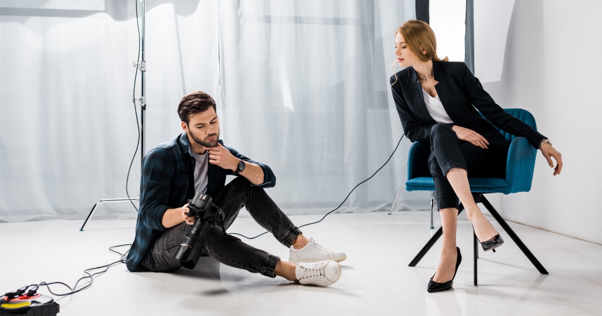 A young professional woman, seated in a chair, looks at a photographer's camera to see the corporate photograph he took of her