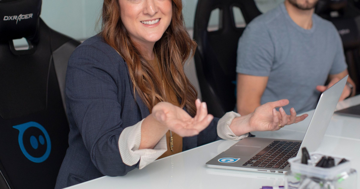 woman with long hair and a blazer shrugs during a conference meeting