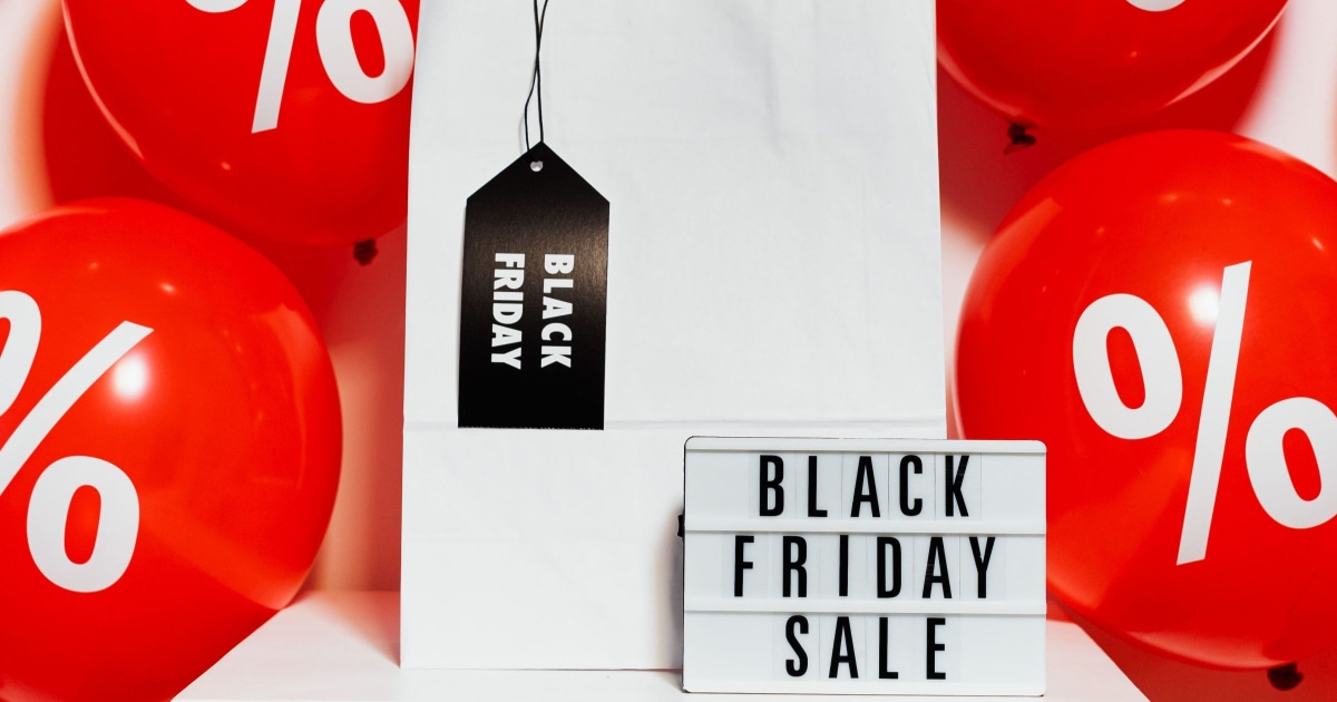 adjustable board reads "BLACK FRIDAY SALE;" it sits in front of a white gift bag with a black tag hanging off it that reads "BLACK FRIDAY." In the background are red balloons with percentage signs.