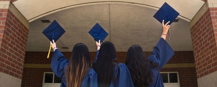 three women in graduation gowns and caps with their backs turned to camera and arms raised