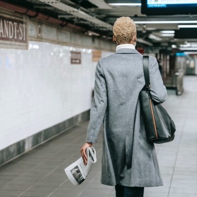 businesswoman carries a tote while walking in the subway; she is Black with bleached hair and wears a gray topcoat, a white blouse, black pants, and her bag is black and brown