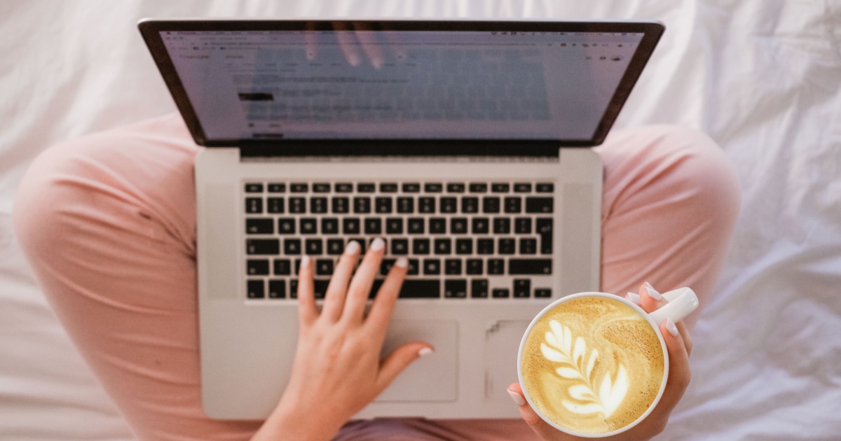 woman wearing pink pants working with laptop on her lap while drinking a fancy coffee