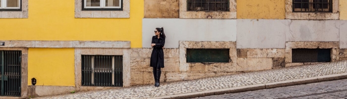 woman stands on cobblestone street; she leans against a wall that is partly yellow