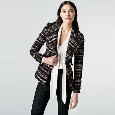 A light-skinned woman with long black hair wearing a black-and-white boucle blazer, white blouse, and black pants
