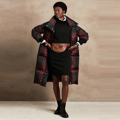 A short-haired Black woman wearing a black dress, a puffy plaid winter coat, and black boots with a brown waist pack