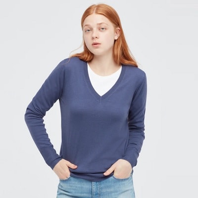 A white woman with long red hair wearing a blue V-neck sweater over a white shirt, and jeans. Her legs are cropped out.
