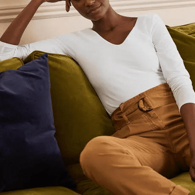 professional woman wears opaque, double-layered v-neck top from Boden; she is lounging on an olive couch next to a navy pillow, and wearing beige pants