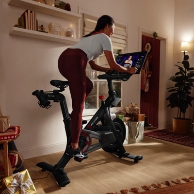 A light-skinned woman riding a Peloton bike in a room with white walls and wooden floor