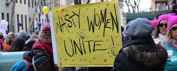 women marching in a crowd, holding a yellow protest sign reading 