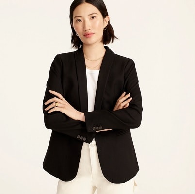 An Asian woman with short black hair wearing a black blazer, white shirt, necklace, and cream-colored pants (legs cropped out)
