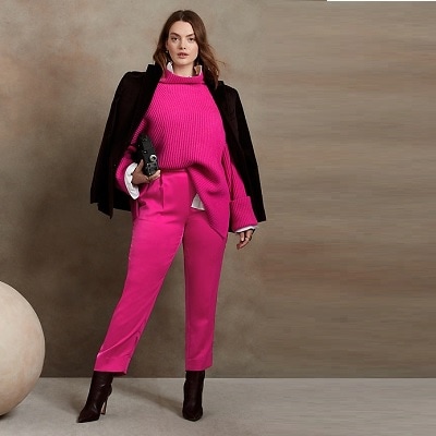 A white woman with long brown hair wearing a pink sweater, black blazer, pink pants, and black high-heeled boots, carrying a black clutch 