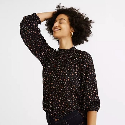 A Black woman with short natural hair wearing a black floral top and black jeans (legs cropped out)
