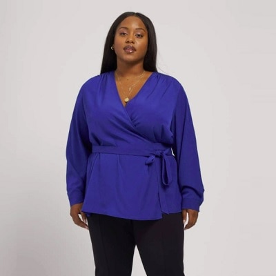 A Black woman with long black hair wearing a blue wrap top, necklace, and black pants (lower legs cropped out)