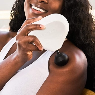 A Black woman with long black hair wearing a white tank top and using a massage gun on her shoulder