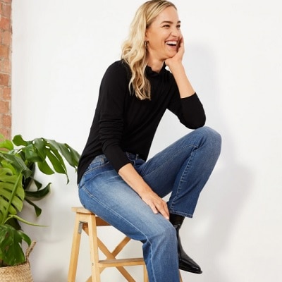 A white woman with long blonde hair wearing a black long-sleeved shirt, jeans, and black boots. She is sitting on  wooden stool and there is a plant in the background.