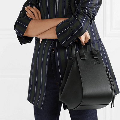 woman in a stripey blazer and jeans stands with her arms crossed, holding a black hammock-shaped leather bag about 12