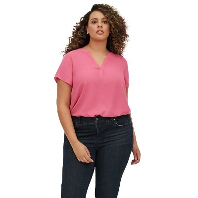 A Black woman with long curly hair wearing a short-sleeved coral top and black pants (lower legs cropped out)