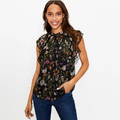A Black woman with long dark brown hair wearing a black floral top and jeans (legs mostly cropped out) 