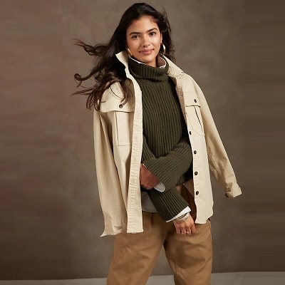A woman with long black hair wearing an olive green sweater with a cream colored utility jacket draped over her shoulders and light brown pants
