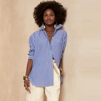 A woman wearing a blue-and-white striped button-front shirt, gold bracelets and necklace, and cream-colored pants (most of legs cropped out)