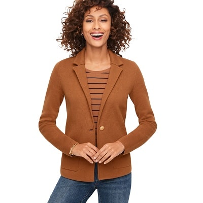 A woman with curly dark brown hair wearing a brown sweater blazer, brown-and-navy striped shirt, and blue jeans (most of legs cropped out)