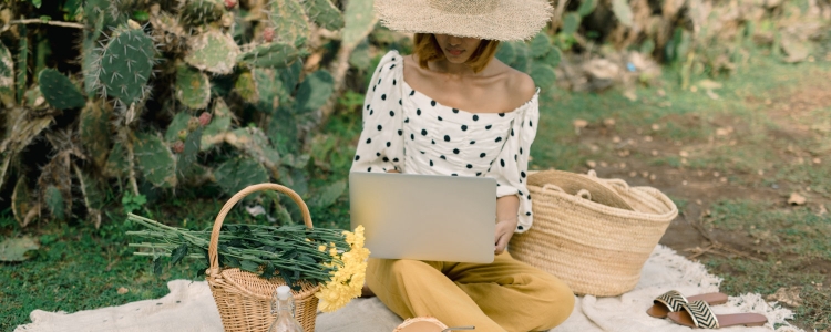 woman wearing a straw hat, an off-the-shoulder white and black polka dot top, and yellow pants sits on a blanket outside with her laptop and some freshly picked flowers