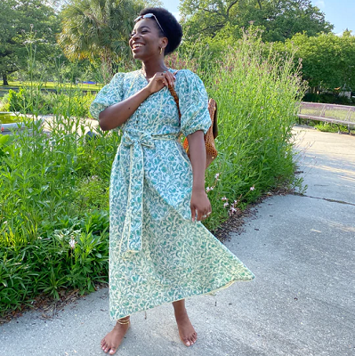 smiling Black woman wearing a beautiful dress with greenish flowers on it; she is barefoot and on a garden path