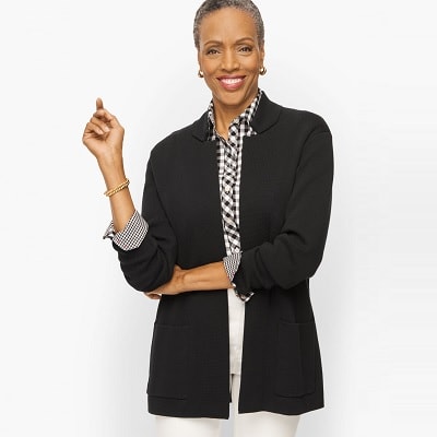 A woman with short black/gray hair wearing a black sweater blazer, a black-and-white gingham shirt, cream-colored pants, and a gold bracelet and earrings