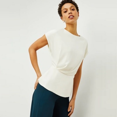 professional woman leans against wall; she wears a short-sleeved white top from M.M.LaFleur with draping details and dark green pants.