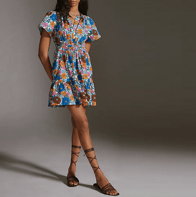 woman in a minidress with smocked waist, short sleeves and a notch neck; the print is orange, blue, and brown flowers against a background of blue leaves. 