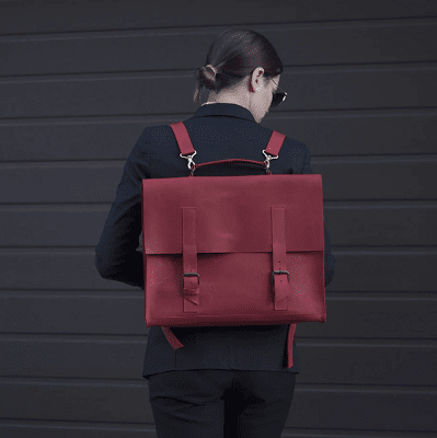 young professional woman has her back turned to the camera; she wears a blood red backpack/satchel/briefcase.