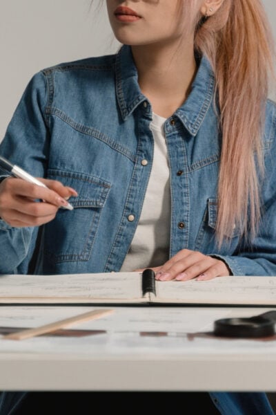 woman with pink and blonde hair works at a desk; she is wearing a blue denim jacket and white t-shirt