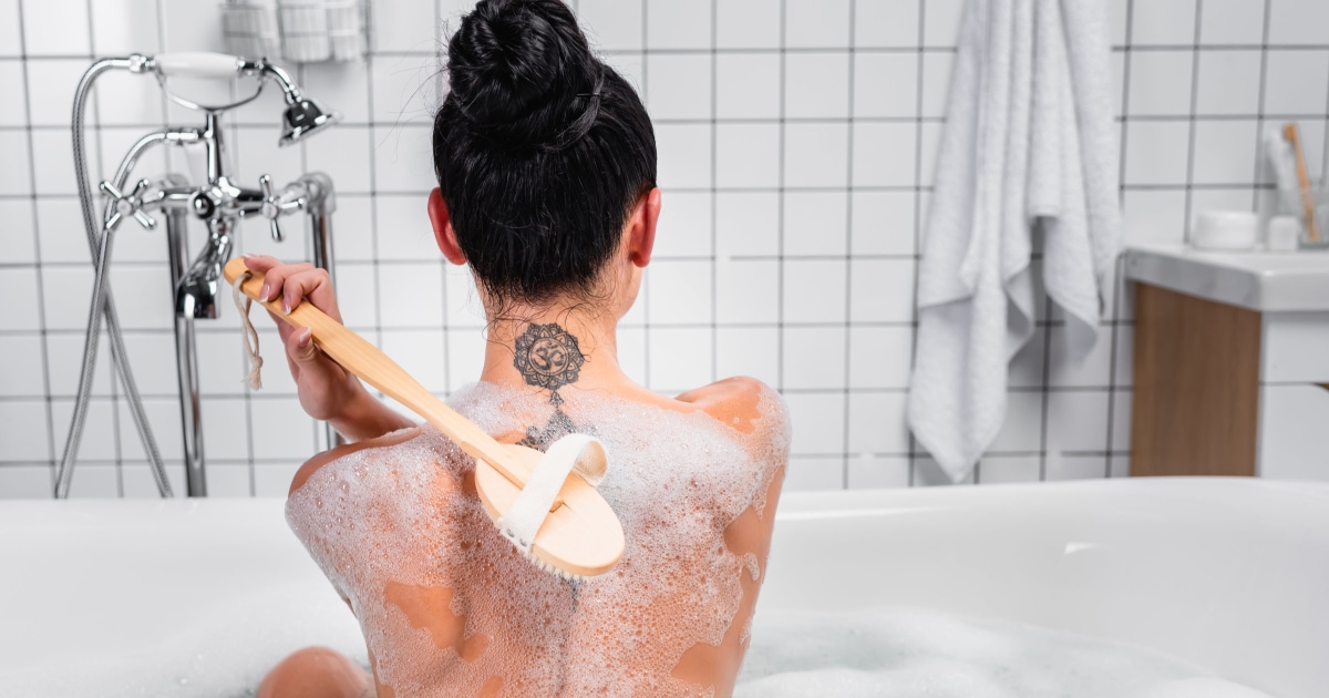 white woman uses a loofah to wash her sudsy back in a bathtub; she has a neck tattoo