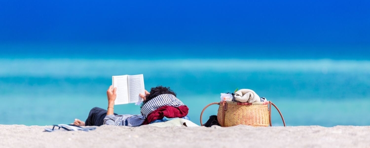 woman lies on beach reading a book, her rattan beach bag sits nearby