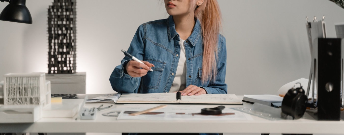 woman with pink and blonde hair works at a desk; she is wearing a blue denim jacket and white t-shirt