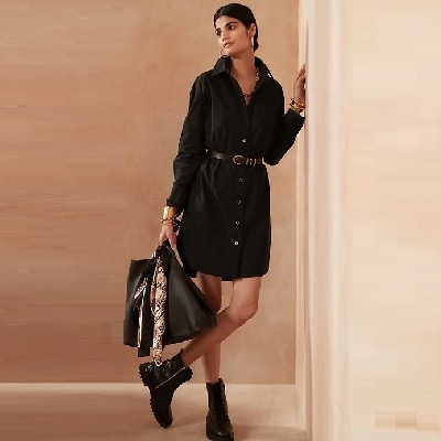 A woman with black hair wearing a black shirtdress, black belt, and black boots, and holding a black bag