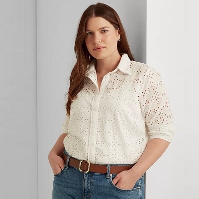 A woman with long brown hair wearing an off-white, button-front eyelet shirt, blue jeans, and a brown belt