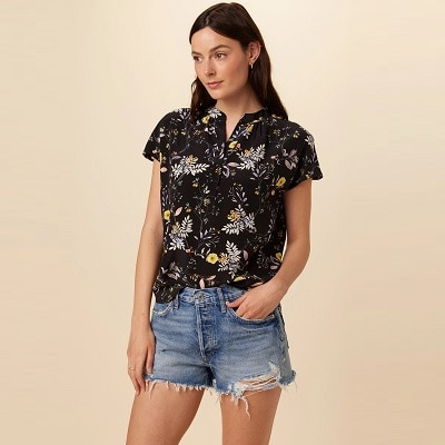 A woman with long brown hair wearing a black floral top and denim cut-off shorts 