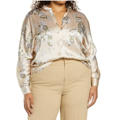 A woman with long black curly hair wearing a cream-colored silk floral top and tan pants