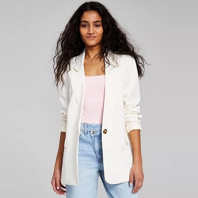 A woman with long black wavy hair wearing a white blazer, pink top, and light blue jeans
