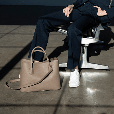 woman slouches in office chair; she wears a suit and bright white sneakers. The grayish brownish tote bag is at her feet. 